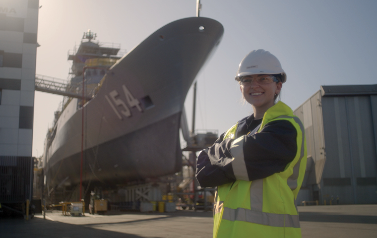 Woman wearing hard hat, safety goggles and hi-vis PPE standing on a shipyard in front of a large ship with number 154 printed on the side.