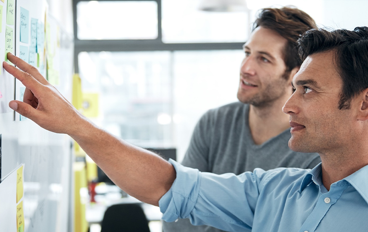 Two men in an office brainstorming using sticky notes on a board. One man is pointing while the other is speaking.