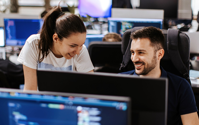 Two people sitting at a desk working on a computer in an office space surrounded by computers.
