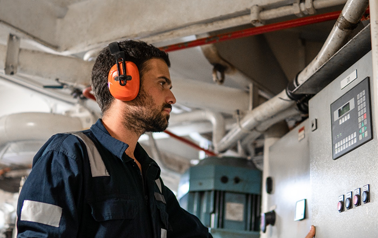 Man wearing protective earwear looking at switchboard around machinery.