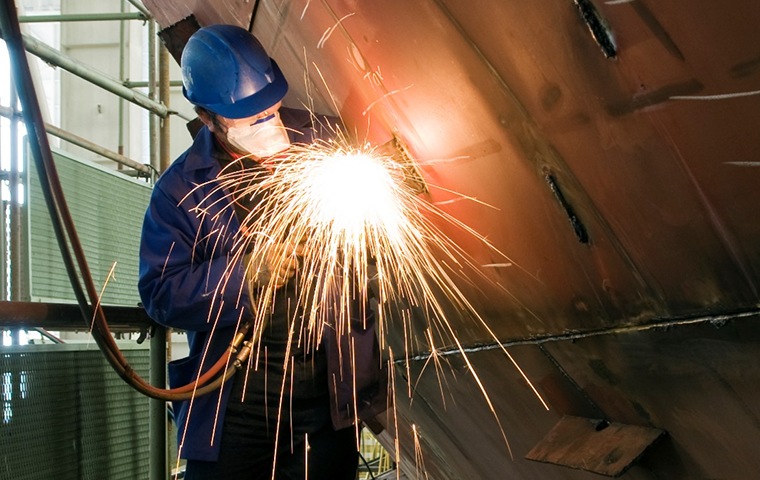 Worker wearing PPE using welding equipment on a ship with sparks flying below them while they weld metal.