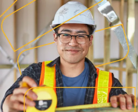 Worker wearing hi vis, hard hat and glasses smiling toward the camera while measuring a beam.