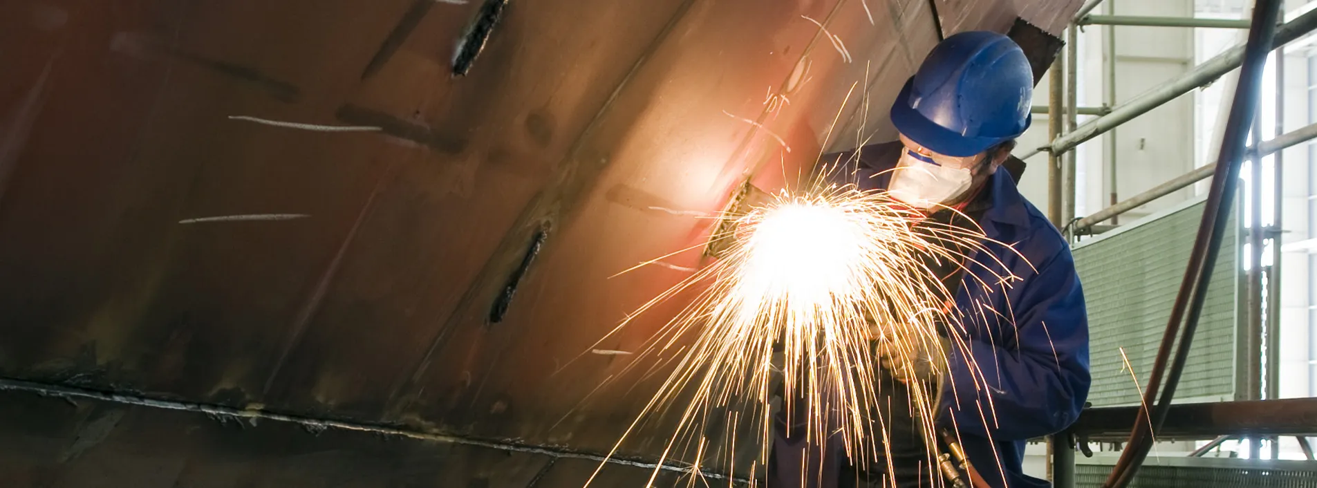 A welder wearing PPE using a welder tool on a ship hull. Sparks are flying.