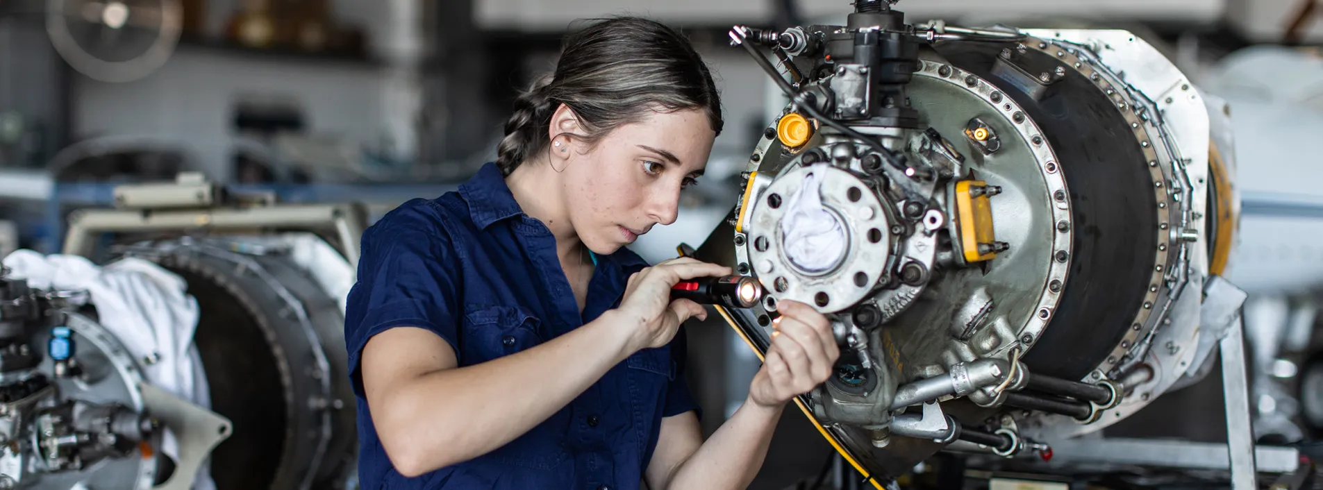 Engineer wearing a blue shirt with her hair tied back inspecting parts of an engine with a small torch