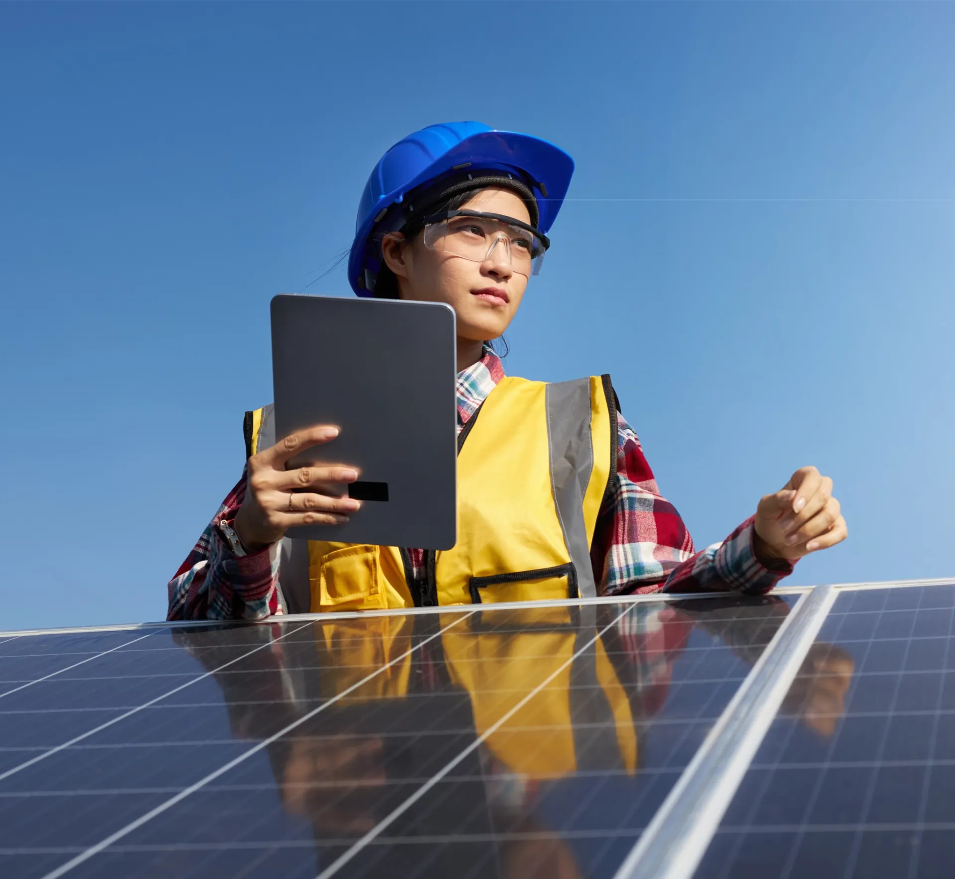 Worker wearing hi vis vest and hard hat while holding a tablet and standing behind a solar panel