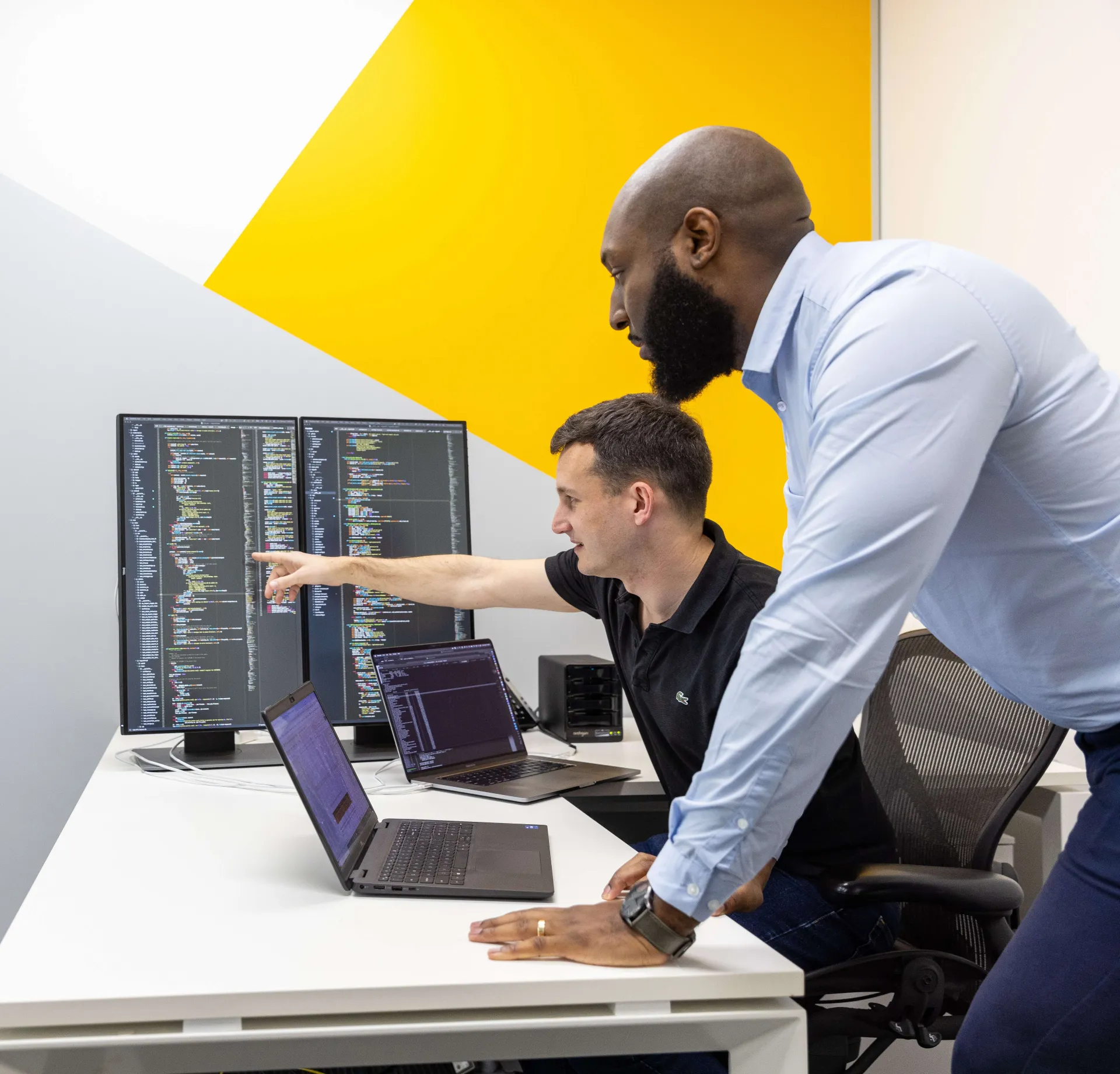 Two IT professionals at a desk inspecting multiple computer and laptop screens that display code