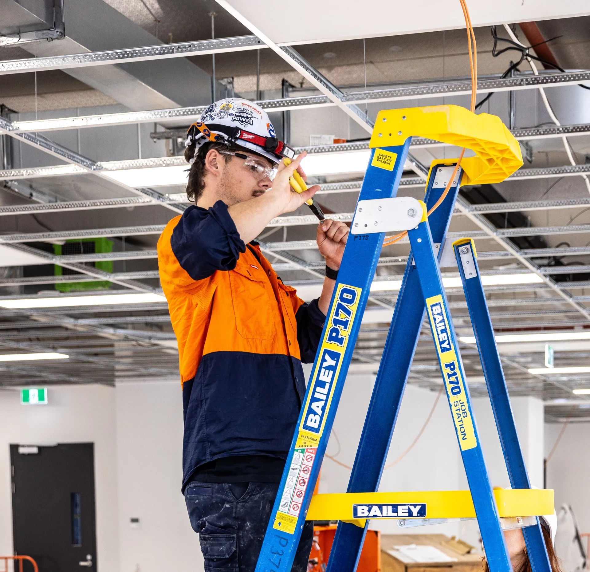 Worker in safety gear using a tool while standing on a ladder inside a construction site.