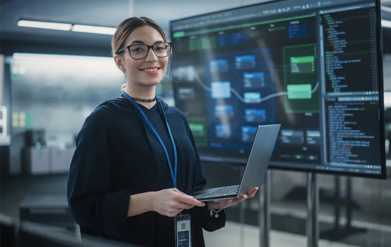 Woman wearing glasses, hoodie and choker, holding a laptop while standing in front of a large screen displaying flowcharts and files.